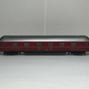 May include: A model train passenger car with a silver roof and a red body. The car has several small windows and the words "SCHLAFWAGEN" and "DSG" printed on the side. The car is on a black track.