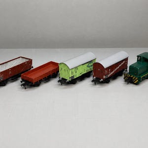 Könnte beinhalten: Eine Sammlung von Modellbahnwagen, darunter ein roter offener Wagen, ein roter Flachwagen, ein grüner gedeckter Wagen, ein weinroter gedeckter Wagen und eine grüne Lokomotive. Die Wagen sind auf einer weißen Oberfläche angeordnet.