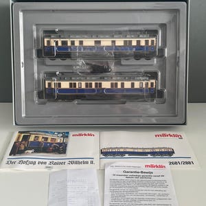 Op de afbeelding: Een modeltreinset met twee crème en blauwe passagierswagons in een grijze koffer. De set bevat gedrukte materialen met de tekst "Der Hofzug von Kaiser Wilhelm II." en "Garantie-Bewijs".