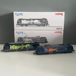 May include: Two Märklin HO scale electric model locomotives displayed in front of their original white packaging boxes. The left locomotive features a black body with green graffiti art, while the right locomotive is blue with a vibrant orange graphic.