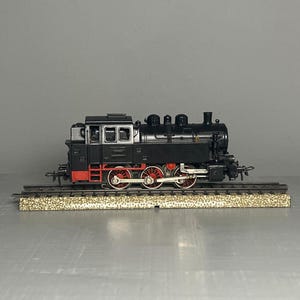 Op de afbeelding: Een zwarte modelstoomlocomotief met rode wielen en zilverkleurige metalen drijfstangen. De trein heeft een gedetailleerde cabine, een cilindrische ketel en een schoorsteen, geplaatst op een kort stuk modelspoor met een textuurrijke basis.