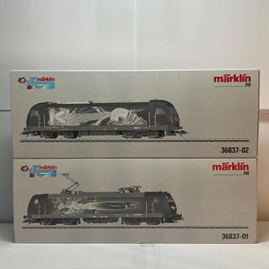 Puede incluir: Dos cajas de trenes modelo Märklin HO. Las cajas son de color gris claro con el logotipo de Märklin e ilustraciones de trenes. Los números de modelo 36837-02 y 36837-01 están impresos en las cajas. Los trenes son negros con diseños detallados.