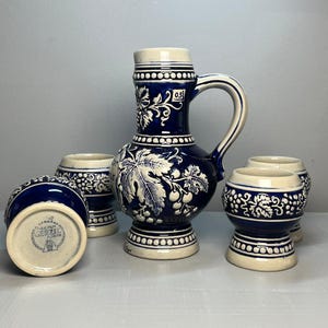 Op de afbeelding: Een blauw-witte keramische kan en vier bijpassende kleine mokken. De kan heeft een handvat en is versierd met druivenblad- en wijnrankontwerpen. De mokken en de kan hebben een crèmekleurige basis en rand.