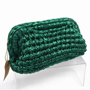 May include: A dark green clutch bag with a woven texture. The bag is rectangular with rounded edges and a small tag attached. The material has a slight sheen, and the bag is displayed on a clear stand.