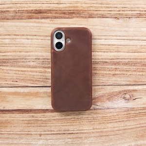 May include: A dark brown leather phone case with a smooth, matte texture. The case features a rectangular shape with rounded corners and a circular cutout at the top left corner, revealing two camera lenses and a small circular flash component.
