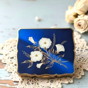 May include: A square, deep blue compact case with a gold-toned metal border. The lid features a decorative inlay of white flowers and delicate, thin gold-coloured stems and leaves arranged in a floral bouquet pattern across the centre surface.