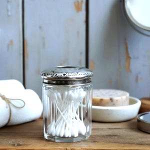 May include: A clear, hexagonal glass jar filled with white cotton buds, topped with a metallic silver lid featuring a decorative perforated edge and a central opening. The container is designed for storage and organisation of small items.