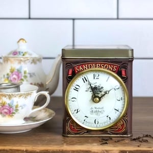 May include: A rectangular metal tin featuring a clock face on the front with the text SANDERSONS at the top. The clock has a white dial with black numerals and hands. The tin has a dark brown finish with decorative red and gold accents.