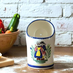 May include: A cylindrical ceramic wall pocket with a slanted top opening, featuring a hand-painted illustration of two figures in traditional attire. The piece is cream-coloured with blue trim and green foliage accents, designed for hanging on a wall.