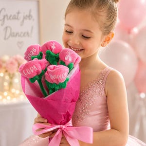Op de afbeelding: Een jong meisje in een roze jurk en tiara houdt een boeket roze stoffen bloemen vast. Elke bloem heeft een groene stengel en een gepersonaliseerde inscriptie. Het boeket is verpakt in roze papier en vastgebonden met een roze lint. Een bord op de achtergrond luidt "Great job, Dancer!"