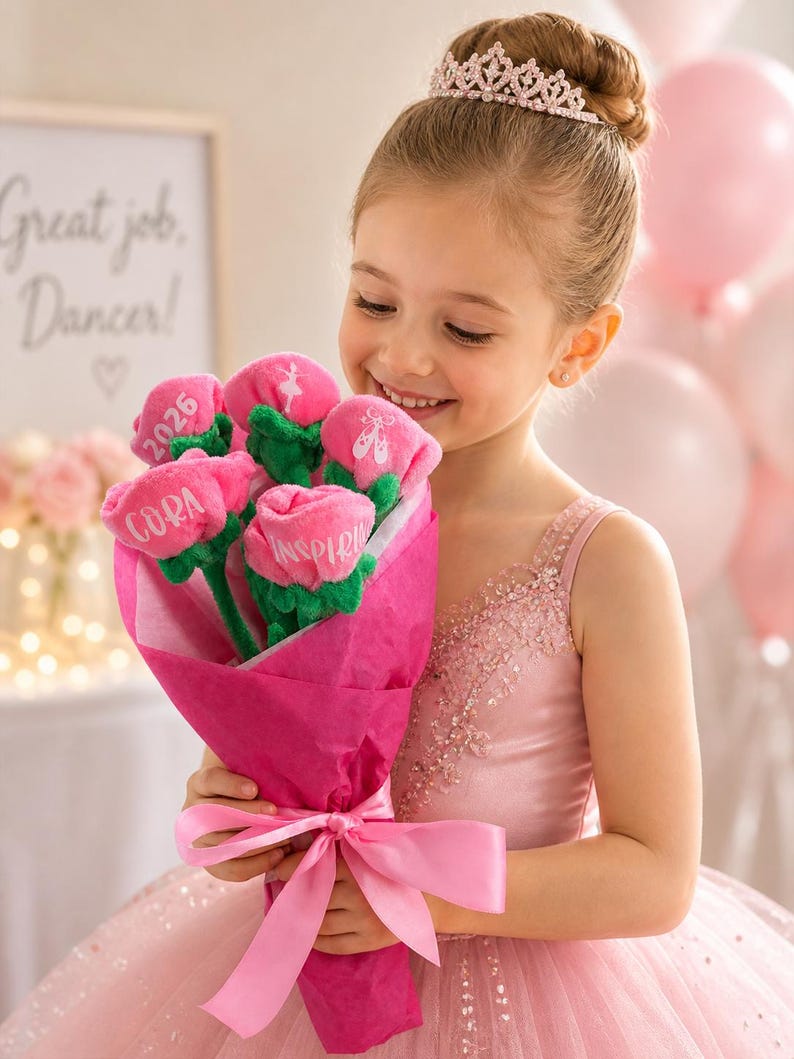 Op de afbeelding: Een jong meisje in een roze jurk en tiara houdt een boeket roze stoffen bloemen vast. Elke bloem heeft een groene stengel en een gepersonaliseerde inscriptie. Het boeket is verpakt in roze papier en vastgebonden met een roze lint. Een bord op de achtergrond luidt "Great job, Dancer!"