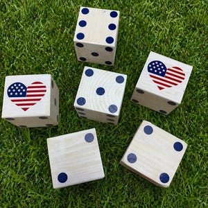 Könnte beinhalten: Sechs Holzwürfel mit blauen Punkten und Herzdesigns mit der amerikanischen Flagge liegen auf grünem Gras verstreut. Die Würfel sind weiß lackiert und haben eine unterschiedliche Anzahl blauer Punkte. Zwei Würfel zeigen ein Herzdesign mit der amerikanischen Flagge.