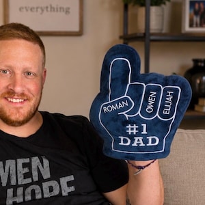 May include: A navy blue foam finger with white trim and the words "#1 DAD." The names "Roman," "Owen," and "Elijah" are printed on the fingers. The foam finger is held up in front of a person.