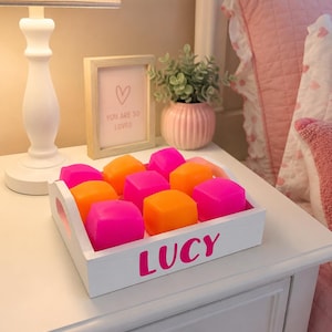May include: A white wooden tray with the name LUCY printed in pink on the front side. The tray contains nine alternating bright neon pink and orange square blocks arranged in a three-by-three grid pattern within the rectangular wooden frame.