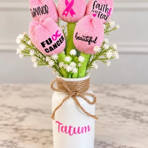 May include: A white ceramic vase with a jute twine bow and the name Tatum in pink text, containing five plush pink tulip-shaped flowers with green stems and white baby's breath. The tulips feature black and pink text: Survivor, a breast cancer ribbon, faith over fear, FU Cancer, and beautiful.