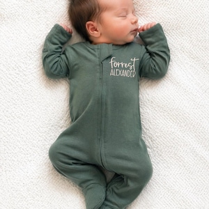 May include: A long-sleeved, footed sleeper in a solid dark sage green color. The garment features a full-length front zipper closure and is personalized with the name "forrest ALEXANDER" embroidered in white script and block lettering on the chest.