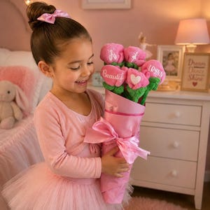 Op de afbeelding: Een jong meisje in een roze balletoutfit houdt een boeket roze pluche bloemen vast. Het boeket is verpakt in roze papier en vastgebonden met een roze lint. Elke bloem heeft een ander ontwerp, waaronder een hart en het woord "Beautiful".