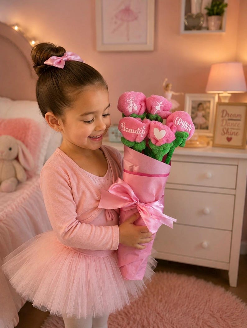 Op de afbeelding: Een jong meisje in een roze balletoutfit houdt een boeket roze pluche bloemen vast. Het boeket is verpakt in roze papier en vastgebonden met een roze lint. Elke bloem heeft een ander ontwerp, waaronder een hart en het woord "Beautiful".