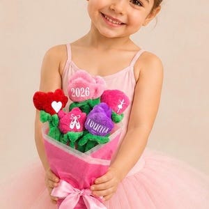 Op de afbeelding: Een jong meisje in een roze turnpakje en tutu houdt een boeket pluche bloemen vast. Het boeket bevat rode, roze en paarse bloemen met tekst en balletthema-ontwerpen. Het boeket is verpakt in roze papier en vastgebonden met een roze lint.