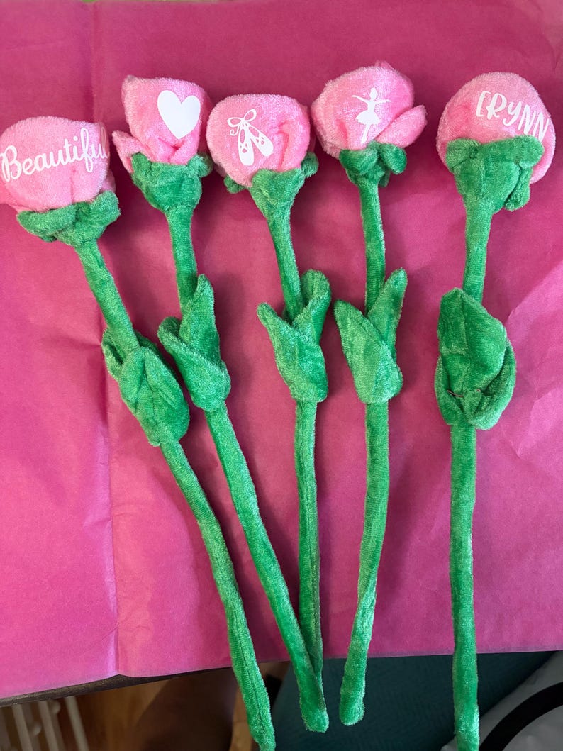 Op de afbeelding: Vijf pluche roze bloemstelen met groene stelen en bladeren. De bloemhoofden hebben witte tekst of ontwerpen, waaronder de woorden "Beautiful" en "RYNN", een hart en balletthema's. De bloemen zijn gerangschikt op een roze oppervlak.