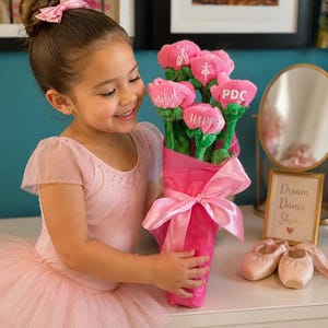 Op de afbeelding: Een jong meisje in een roze balletoutfit houdt een boeket roze pluche bloemen vast. De bloemen zijn verpakt in roze papier en vastgebonden met een roze lint. De bloemen hebben de woorden "Beautiful" en "Happy".