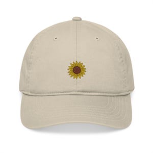 May include: A beige baseball cap with a sunflower embroidered on the front. The sunflower has yellow petals and a brown center. The cap has a curved brim and a six-panel design.