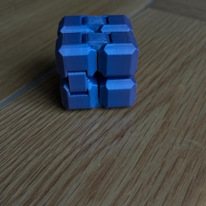 May include: A blue fidget cube with a geometric design. The cube is made of several connected blocks that can be rotated and manipulated. The cube is sitting on a wooden surface.