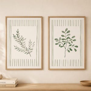 Puede incluir: Dos cuadros rectangulares con marcos de madera clara, cada uno con una ilustración botánica verde de ramas con hojas centrada, enmarcada por bordes de rayas verticales sobre un fondo crema, expuestos lado a lado en una pared.
