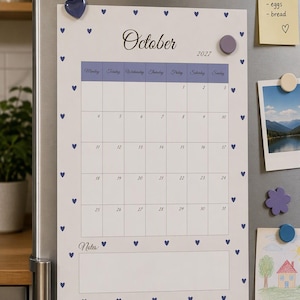 Puede incluir: Un calendario de pared blanco para octubre de 2027 con un encabezado morado y un borde de pequeños corazones azules. El calendario presenta una cuadrícula para las fechas, una sección de notas en la parte inferior y un imán de corazón.
