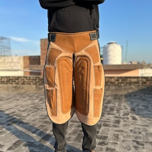 May include: A pair of brown leather farrier chaps with pockets and a black belt. The chaps are worn over black jeans and brown leather shoes. The chaps have a lighter brown padded area on the front.