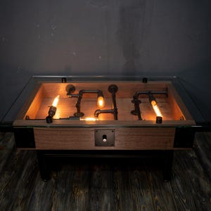 Könnte beinhalten: Einzigartiger Couchtisch mit Glasplatte, Holzrahmen und schwarzen Metallbeinen. Im Inneren befindet sich eine Holzkiste mit dekorativen schwarzen Rohren und beleuchteten Glühbirnen. Der Tisch steht auf einem dunklen Holzboden.