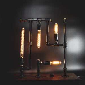 May include: Industrial-style lamp featuring black metal pipes and fittings, with Edison-style light bulbs. The lamp is mounted on a dark wooden base. The bulbs emit a warm, inviting glow against a dark background.