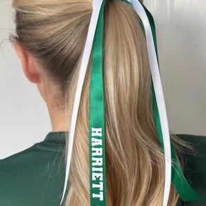 May include: A hair accessory featuring two long satin ribbons in solid white and emerald green tied around a ponytail. The green ribbon displays the name HARRIETT printed in bold white capital letters. The ribbons hang down freely.