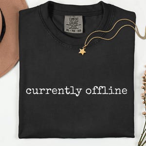 May include: Black crew neck t-shirt with the words "currently offline" in white. A gold necklace with a star pendant rests on the shirt. The shirt has a Comfort Colors tag.