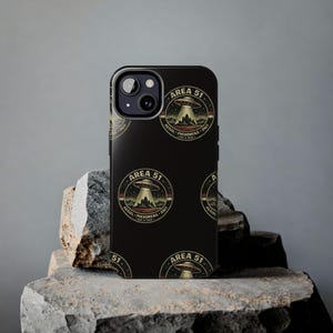 May include: Black phone case featuring a repeating circular design with the text "AREA 51" and a graphic of a UFO over a mountain. The design also includes the words "AERIAL PHENOMENA UNIT". The case is displayed on a gray stone surface.
