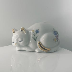 May include: A white ceramic cat figurine in a sleeping pose, with gold accents on the ears and tail. The cat is decorated with floral patterns in blue, yellow, and purple. The figurine is on a white surface.