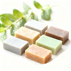 Può includere: Un assortimento di saponi rettangolari in vari colori, tra cui beige, verde e marrone, disposti su una superficie bianca. Foglie verdi sullo sfondo. I saponi sembrano fatti a mano e naturali.