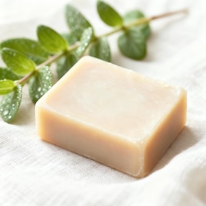 May include: A rectangular bar of light beige soap rests on a white, textured cloth. Fresh green leaves with water droplets are in the background. The soap has a smooth surface and rounded edges, suggesting a natural, handmade product.