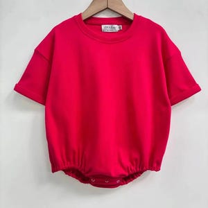 May include: A bright red baby romper with short sleeves and a round neckline. The romper has a gathered waist and snaps at the bottom. The label reads "LEVI + LUNA". The garment is hanging on a wooden hanger.