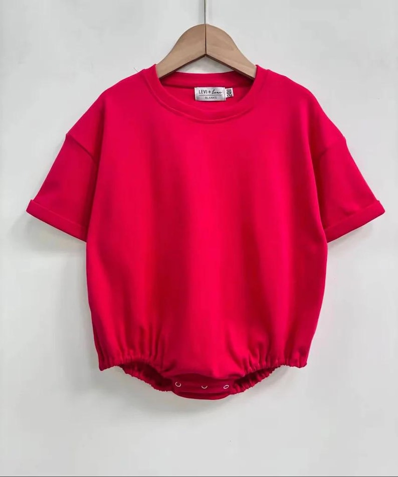 May include: A bright red baby romper with short sleeves and a round neckline. The romper has a gathered waist and snaps at the bottom. The label reads "LEVI + LUNA". The garment is hanging on a wooden hanger.