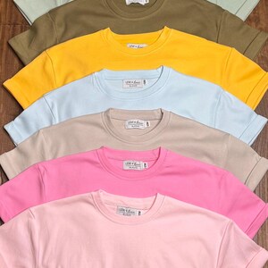 May include: A collection of short-sleeved t-shirts and a romper in various pastel colors, including white, tan, lavender, green, olive, yellow, blue, beige, pink, and light pink. Each shirt has a crew neck and a small tag. The romper is light pink.