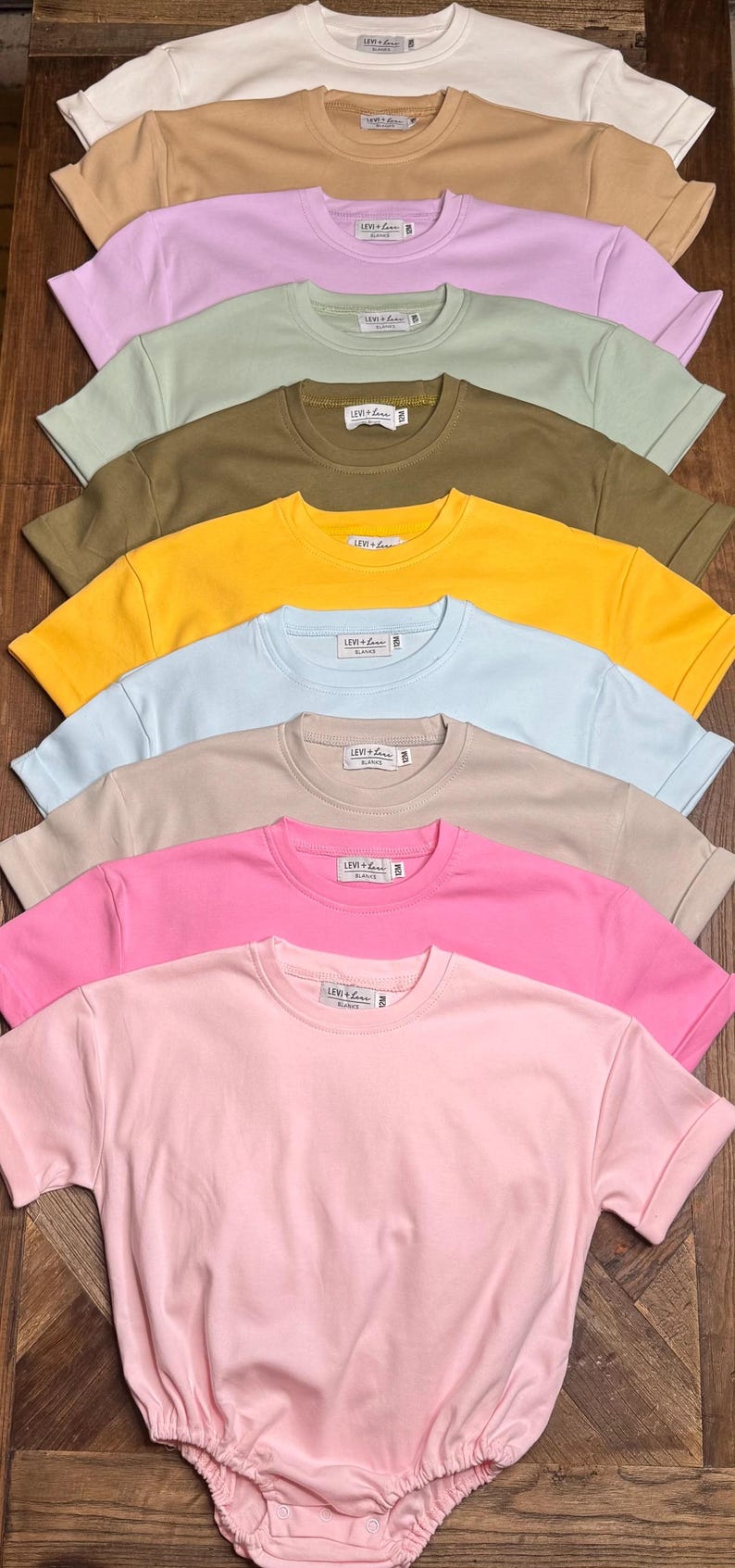 May include: A collection of short-sleeved t-shirts and a romper in various pastel colors, including white, tan, lavender, green, olive, yellow, blue, beige, pink, and light pink. Each shirt has a crew neck and a small tag. The romper is light pink.