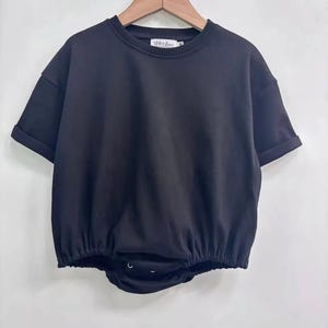 May include: Black short-sleeved baby bodysuit with a round neckline and rolled sleeves. The bottom portion is gathered with a snap closure. The brand name "LEVI + LOU" is printed on the neckline.