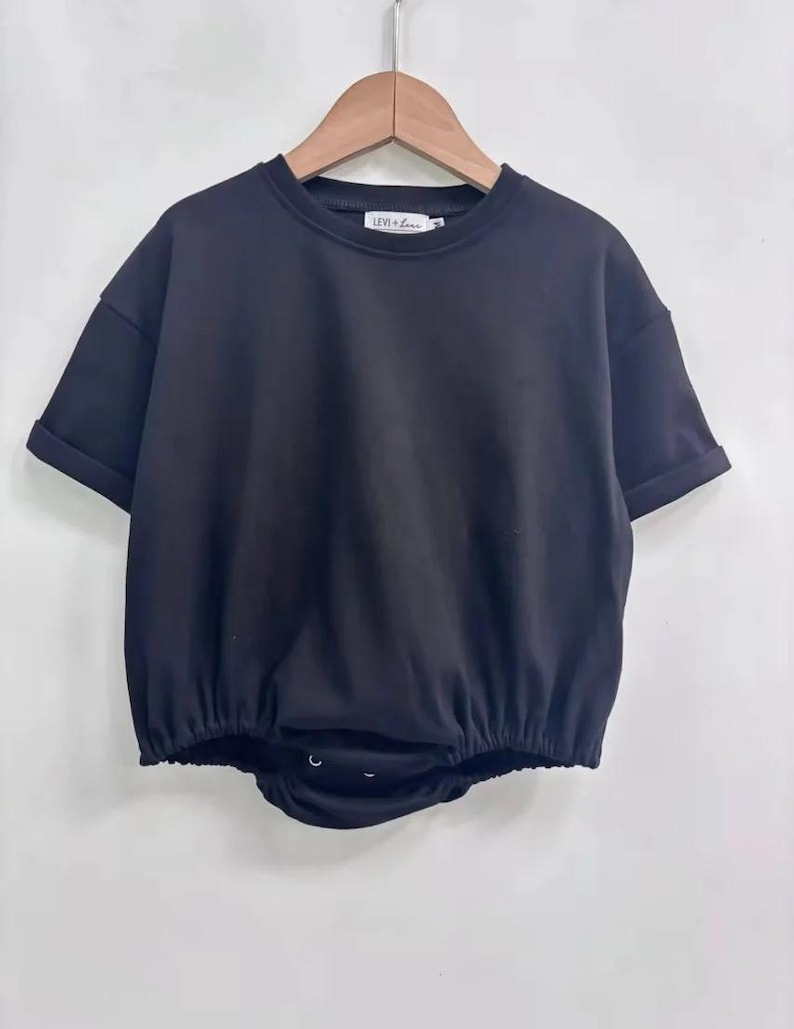 May include: Black short-sleeved baby bodysuit with a round neckline and rolled sleeves. The bottom portion is gathered with a snap closure. The brand name "LEVI + LOU" is printed on the neckline.