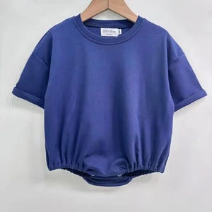 May include: A solid blue baby romper with short sleeves and a round neckline. The romper has a gathered waist and snaps at the bottom. The brand name "LEVI + LOU" is printed on a small tag at the neckline. The garment is hanging on a wooden hanger.