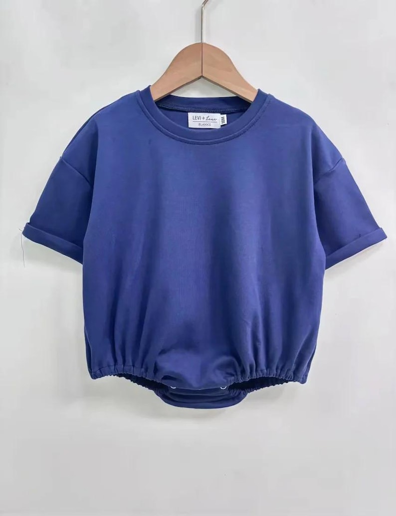 May include: A solid blue baby romper with short sleeves and a round neckline. The romper has a gathered waist and snaps at the bottom. The brand name "LEVI + LOU" is printed on a small tag at the neckline. The garment is hanging on a wooden hanger.