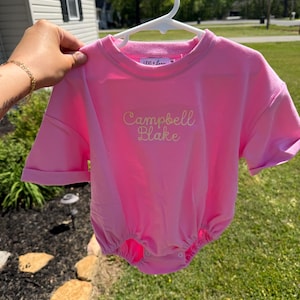 May include: A bright pink baby romper with short sleeves and a round neckline. The romper has the embroidered text "Campbell Blake" in a cursive font. The brand name "LEVI + LOU" is embroidered on the neckline.
