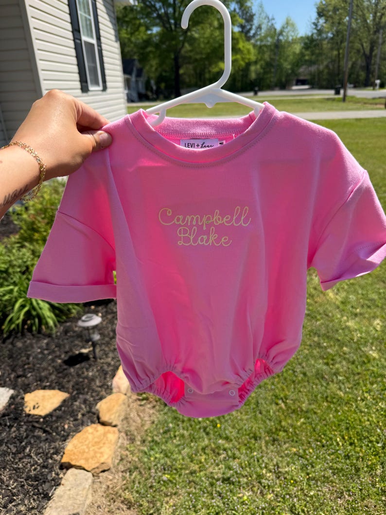 May include: A bright pink baby romper with short sleeves and a round neckline. The romper has the embroidered text "Campbell Blake" in a cursive font. The brand name "LEVI + LOU" is embroidered on the neckline.