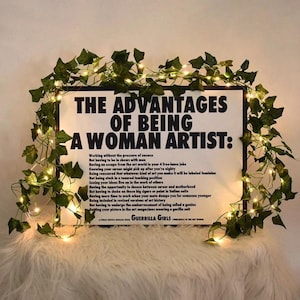 May include: A framed poster with the text "THE ADVANTAGES OF BEING A WOMAN ARTIST:" and a list of advantages. The poster is decorated with green vines and fairy lights. The text "GUERRILLA GIRLS" is at the bottom.