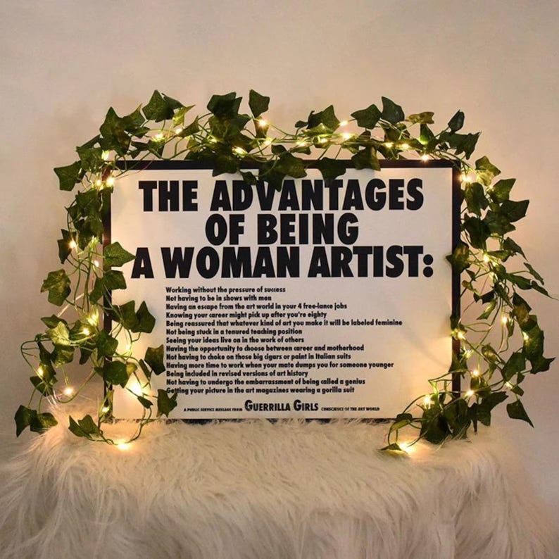 May include: A framed poster with the text "THE ADVANTAGES OF BEING A WOMAN ARTIST:" and a list of advantages. The poster is decorated with green vines and fairy lights. The text "GUERRILLA GIRLS" is at the bottom.
